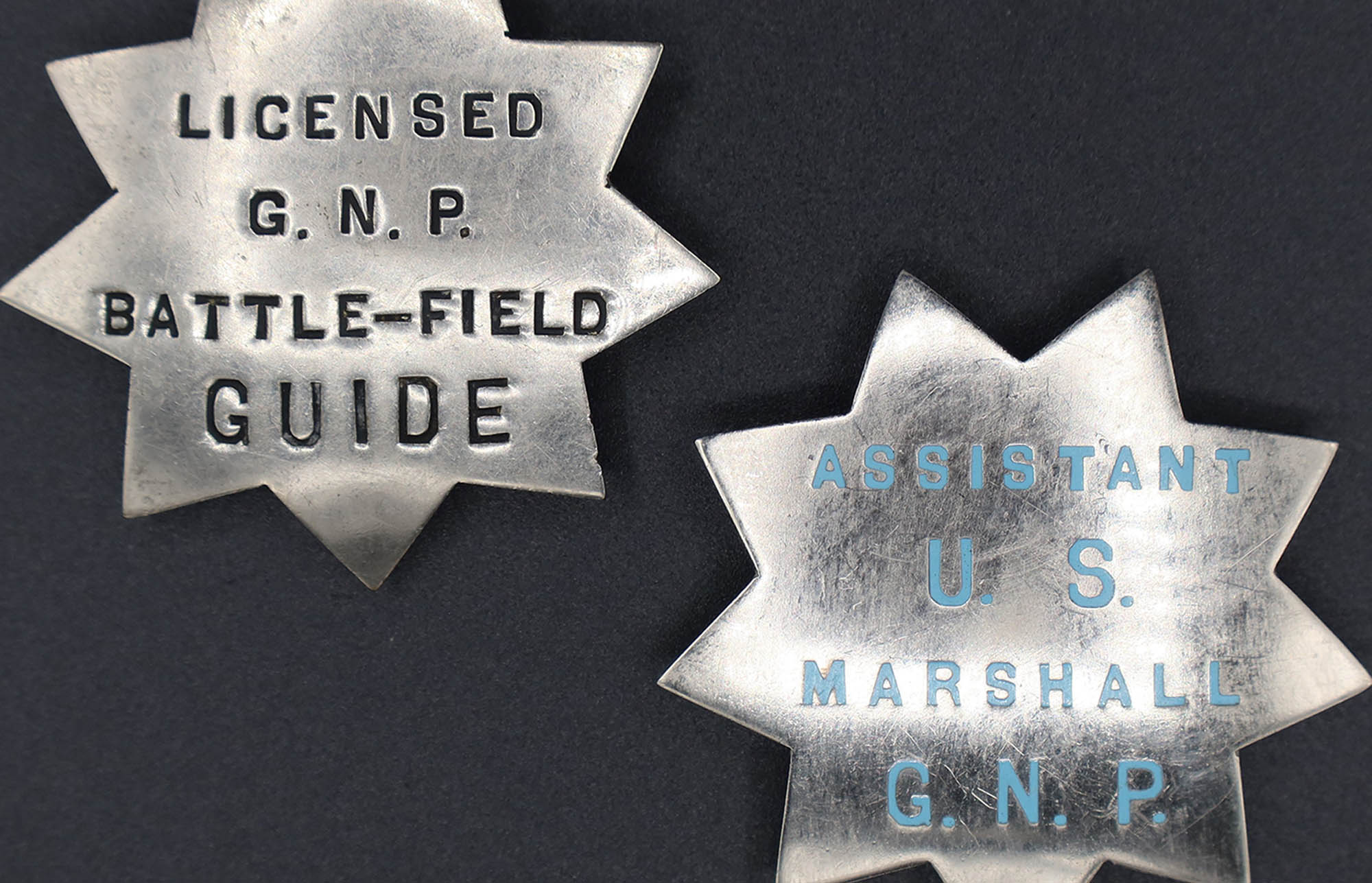Two silver badges shaped as 9-point stars. One is marked with black letters for a licensed guide. The other in blue for an assistant marshal.